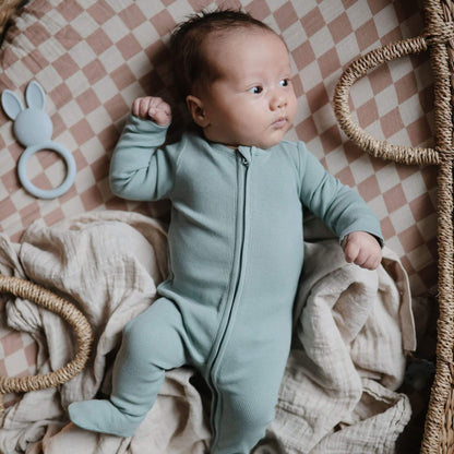 Baby in Mushie Zipper Pajama - Gren lying on a checkered blanket with toys around