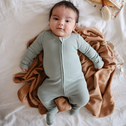 A baby lying on a white bed, wearing Mushie Zipper Pajama - Green and wrapped in a brown blanket.