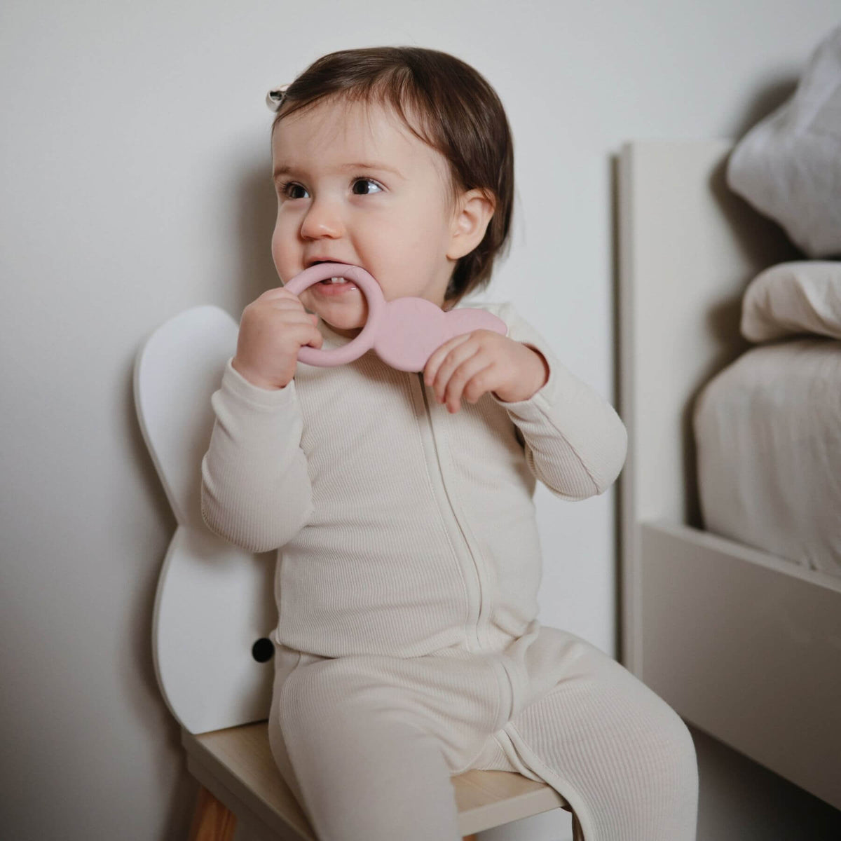 Baby sitting on a chair holding a pink teething ring.