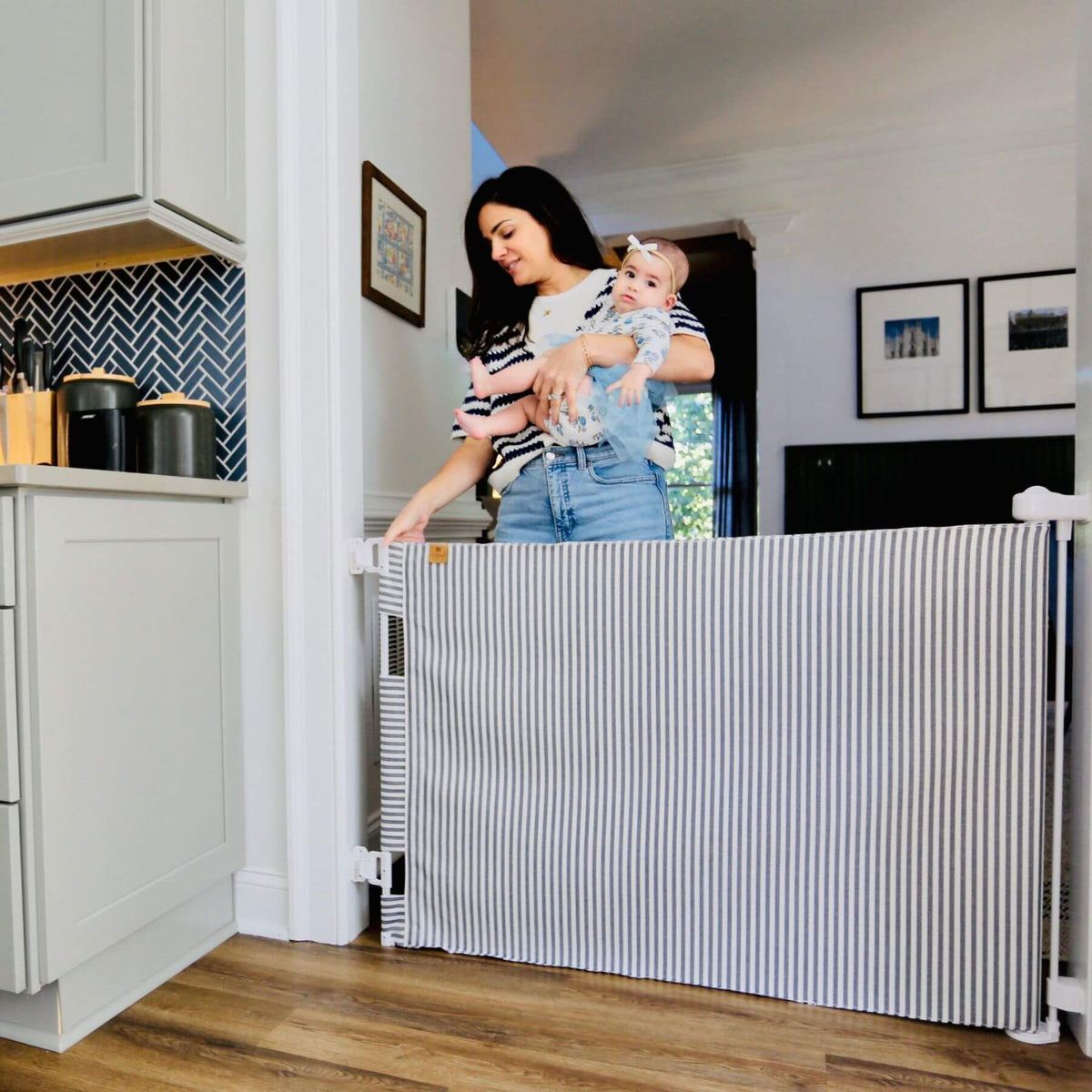 A woman holding a baby stands behind a striped baby safety gate in a kitchen doorway. The gate has a white top and bottom frame with narrow vertical gray and white stripes across the panels.