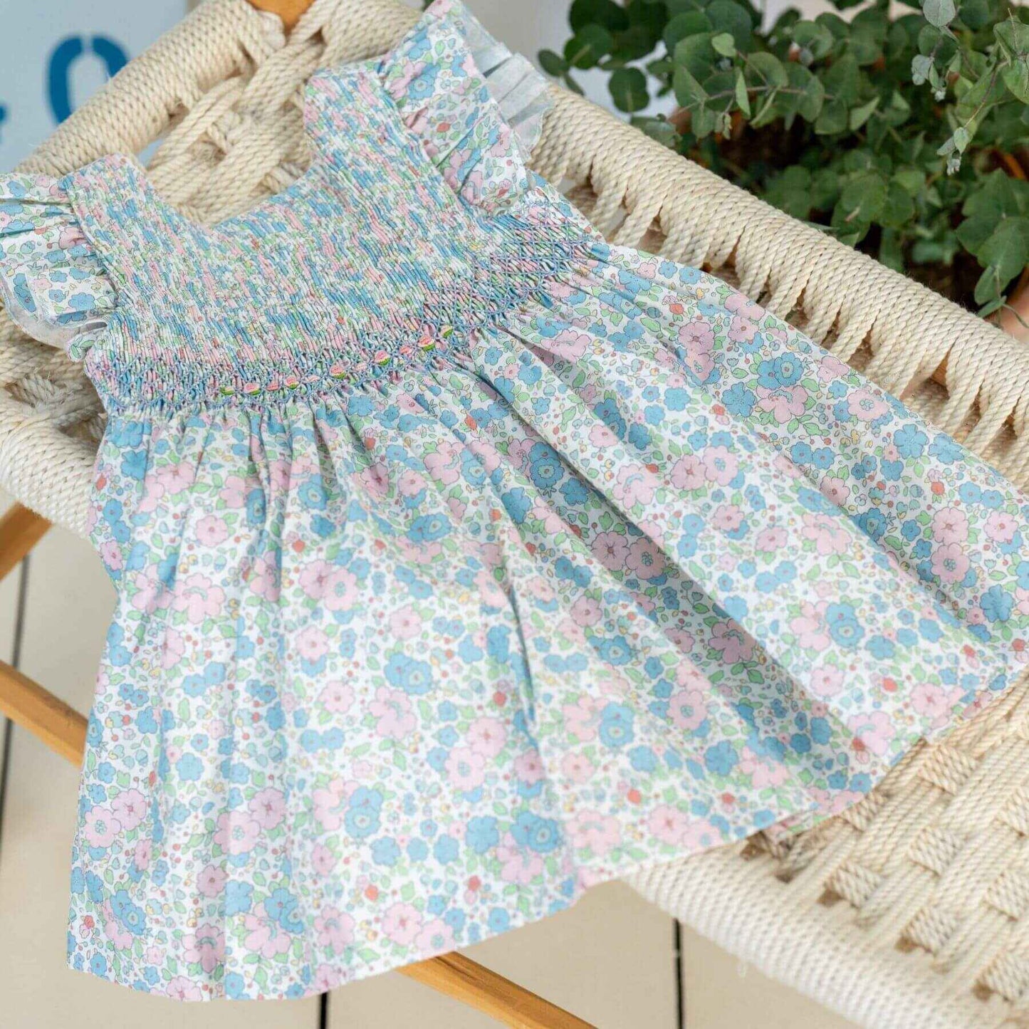 NELIA | Girls Blue Floral Cotton Smocked Dress on a chair with a plant in the background