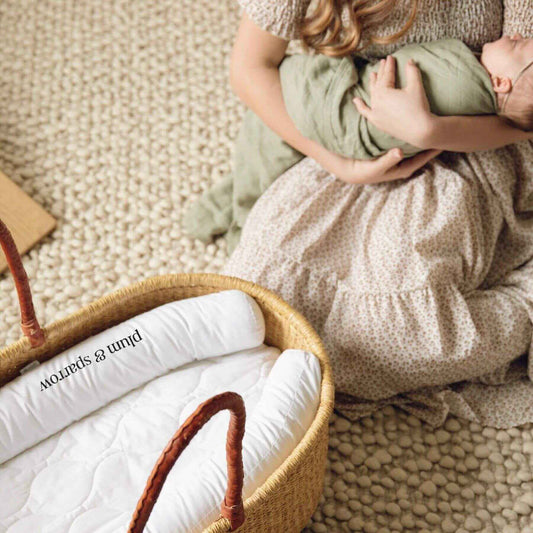 Person holding a baby wrapped in a green blanket next to Plum+Sparrow Nest Lounger.