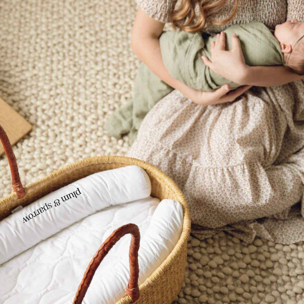 Person holding a baby wrapped in a green blanket next to Plum+Sparrow Nest Lounger.