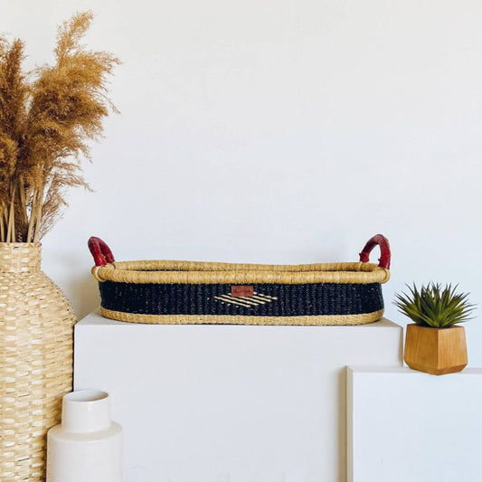 Plum+Sparrow Nightfall Changing Basket on a white surface, next to a vase with dried plants and a small potted plant.