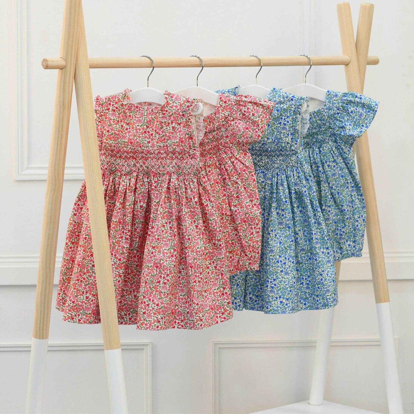 Four floral dresses, two red and two blue, hanging on a wooden rack against a white wall.