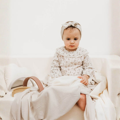 Baby in a floral outfit sitting on Little Mae & Co Oat Ribbed Toddler Blanket