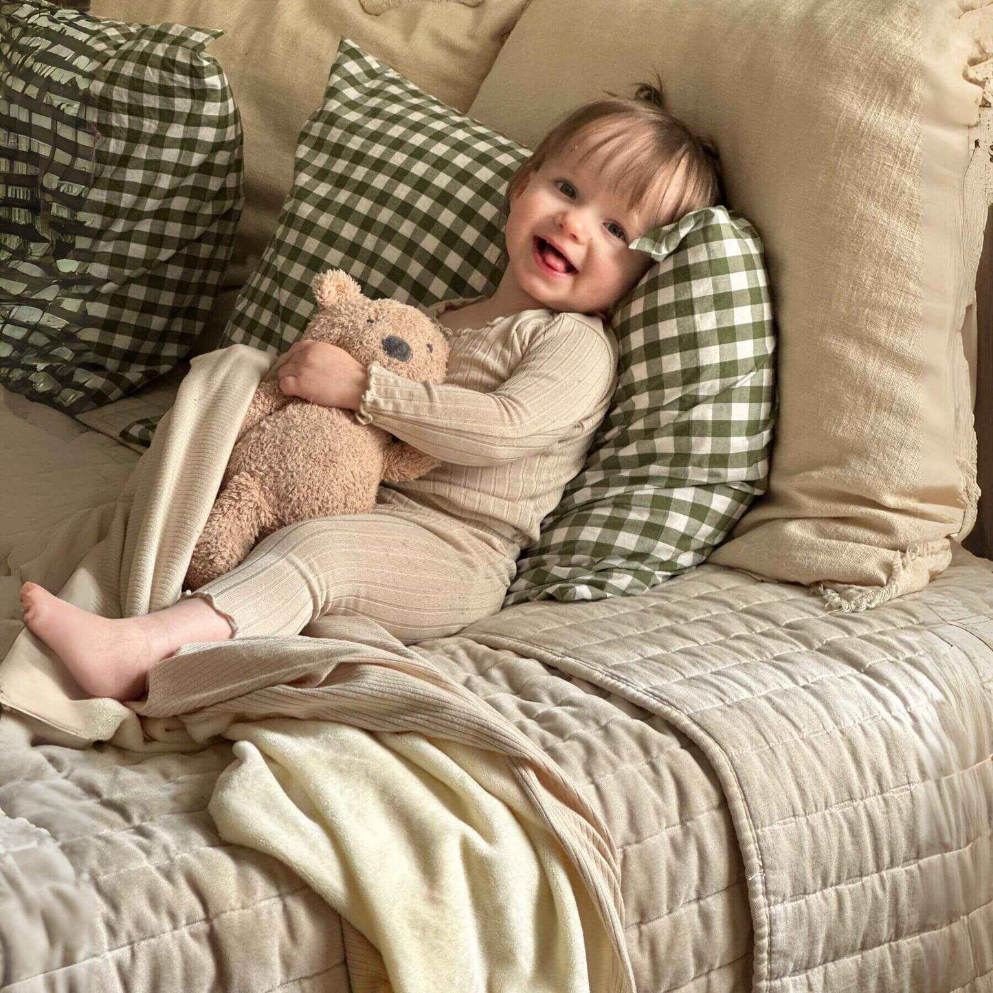 Child sitting on Little Mae & Co Oat Ribbed Toddler Blanket holding a teddy bear with green checkered pillows.