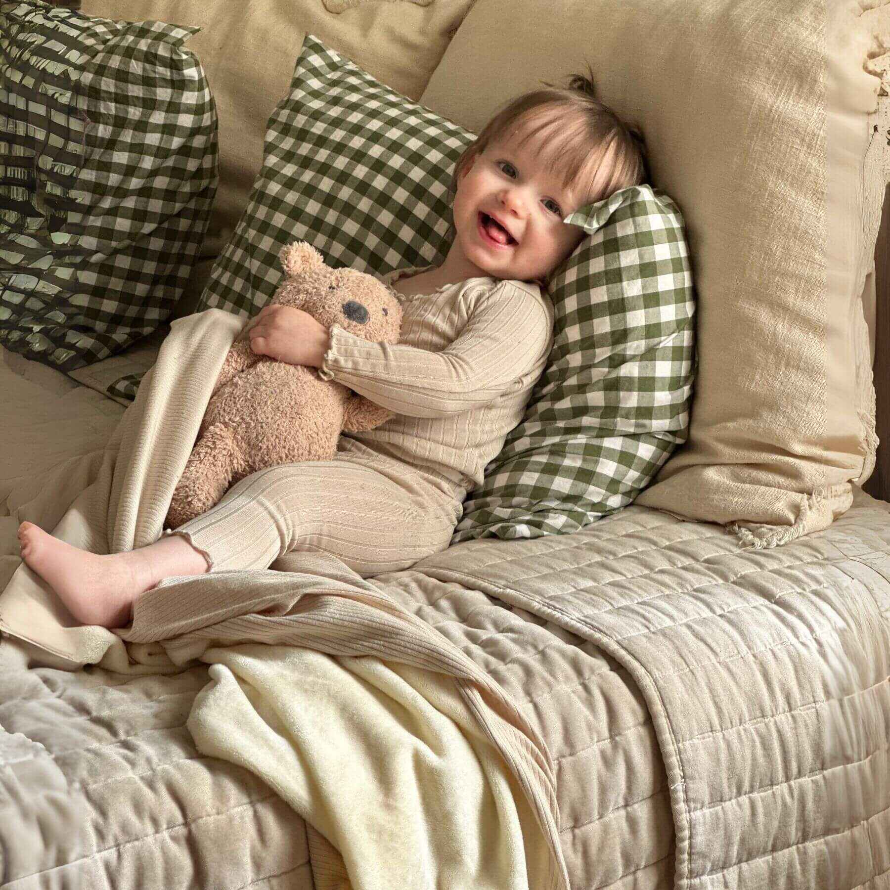 Child sitting on Little Mae & Co Oat Ribbed Toddler Blanket holding a teddy bear with green checkered pillows.
