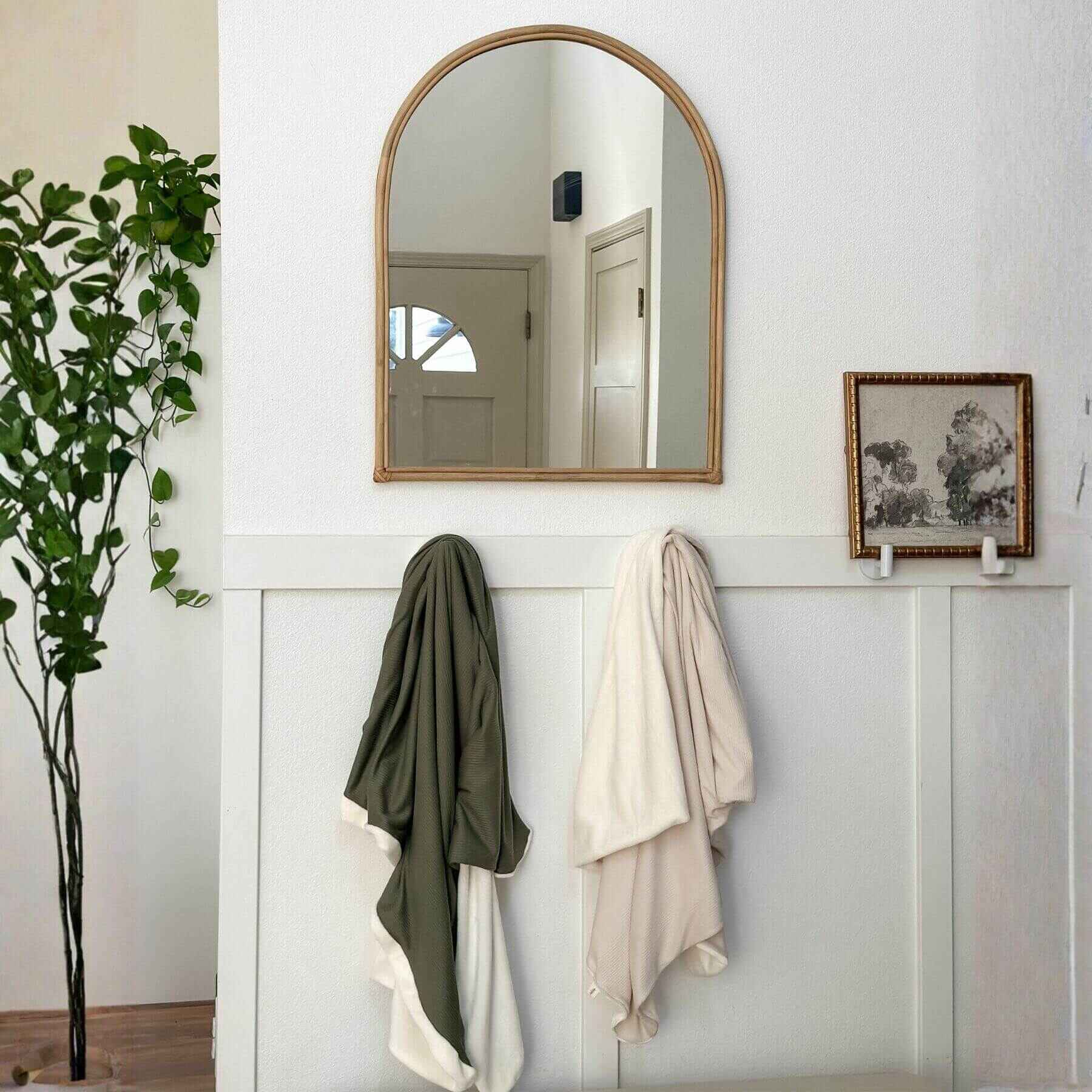 Wooden mirror on a white wall with two cloths hanging below it, a plant to the left, and a framed picture on the right.