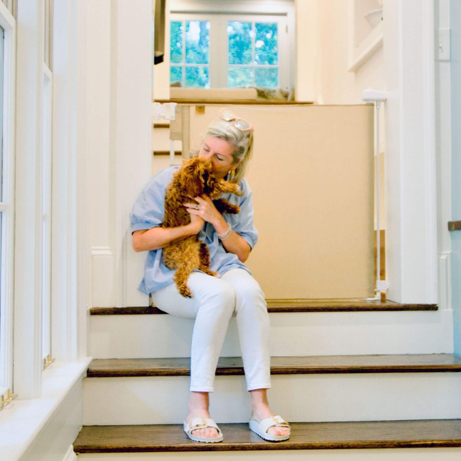Woman sitting on a staircase holding a dog, with Crafted Gates Co. Oil Waxed Canvas Pet Safety Gate - Tan and window in the background.