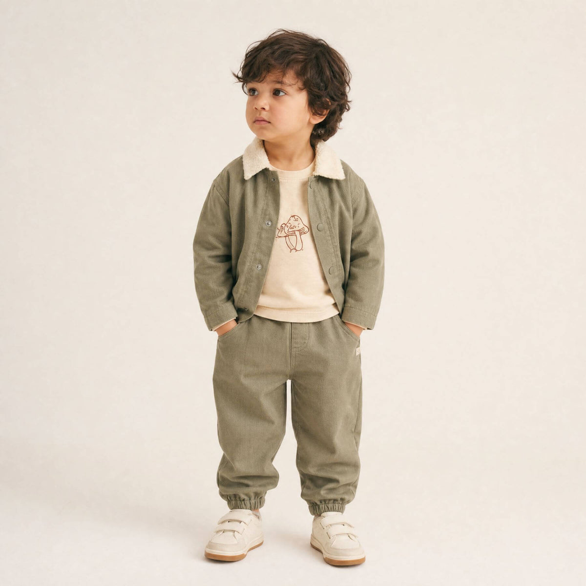 A toddler stands against a plain light beige studio background wearing a matching two-piece outfit consisting of an olive green corduroy-style jacket with a cream collar and matching corduroy pants.