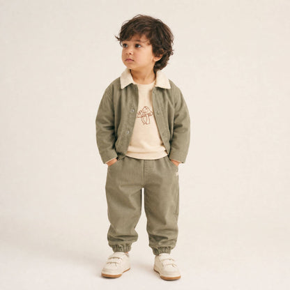 A toddler stands against a plain light beige studio background wearing a matching two-piece outfit consisting of an olive green corduroy-style jacket with a cream collar and matching corduroy pants.