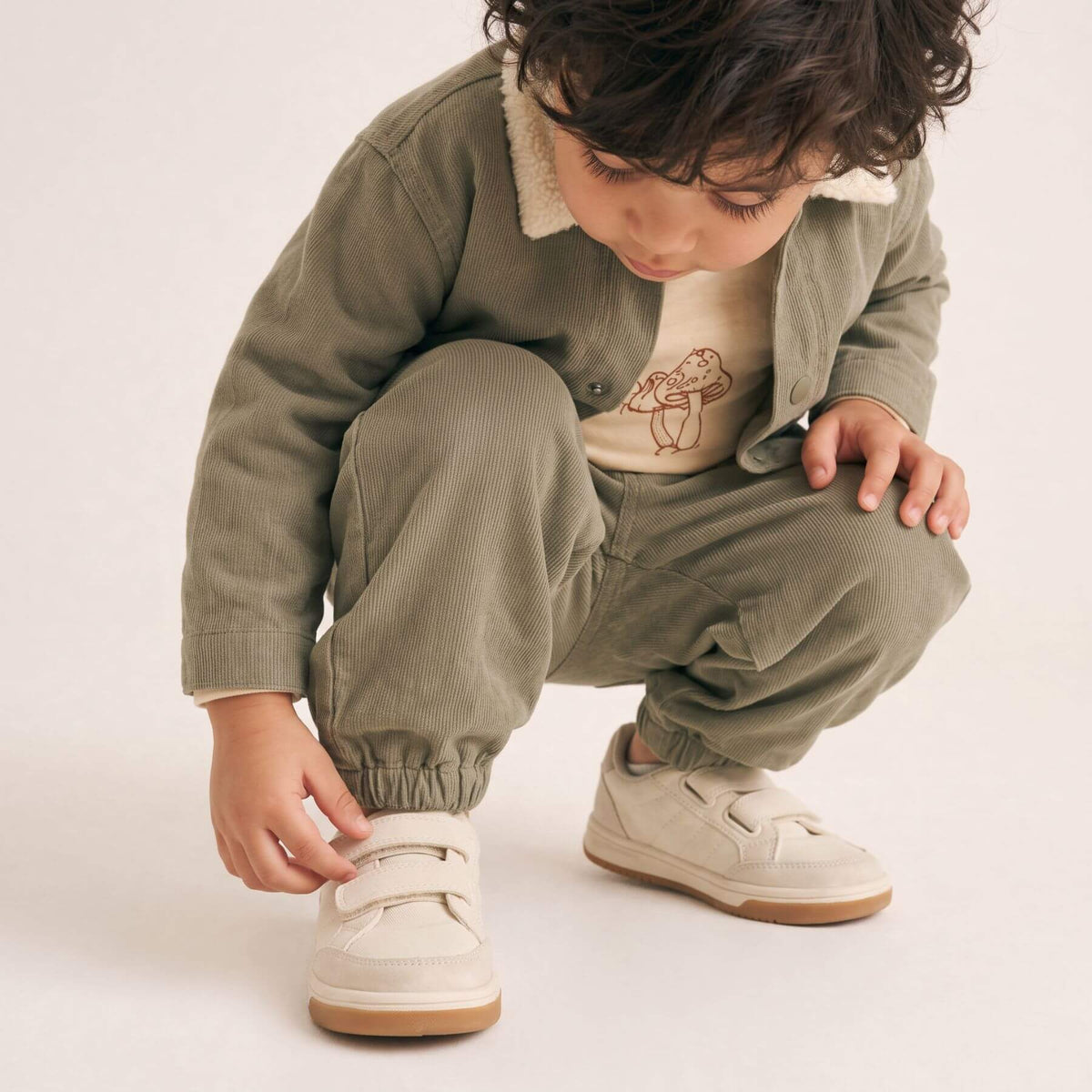 A toddler crouching down to tie the velcro strap of their cream-colored low-top sneakers. The child is wearing an olive green corduroy jacket with a cream sherpa collar and matching corduroy trousers, layered over a beige t-shirt with a teddy bear graphic.