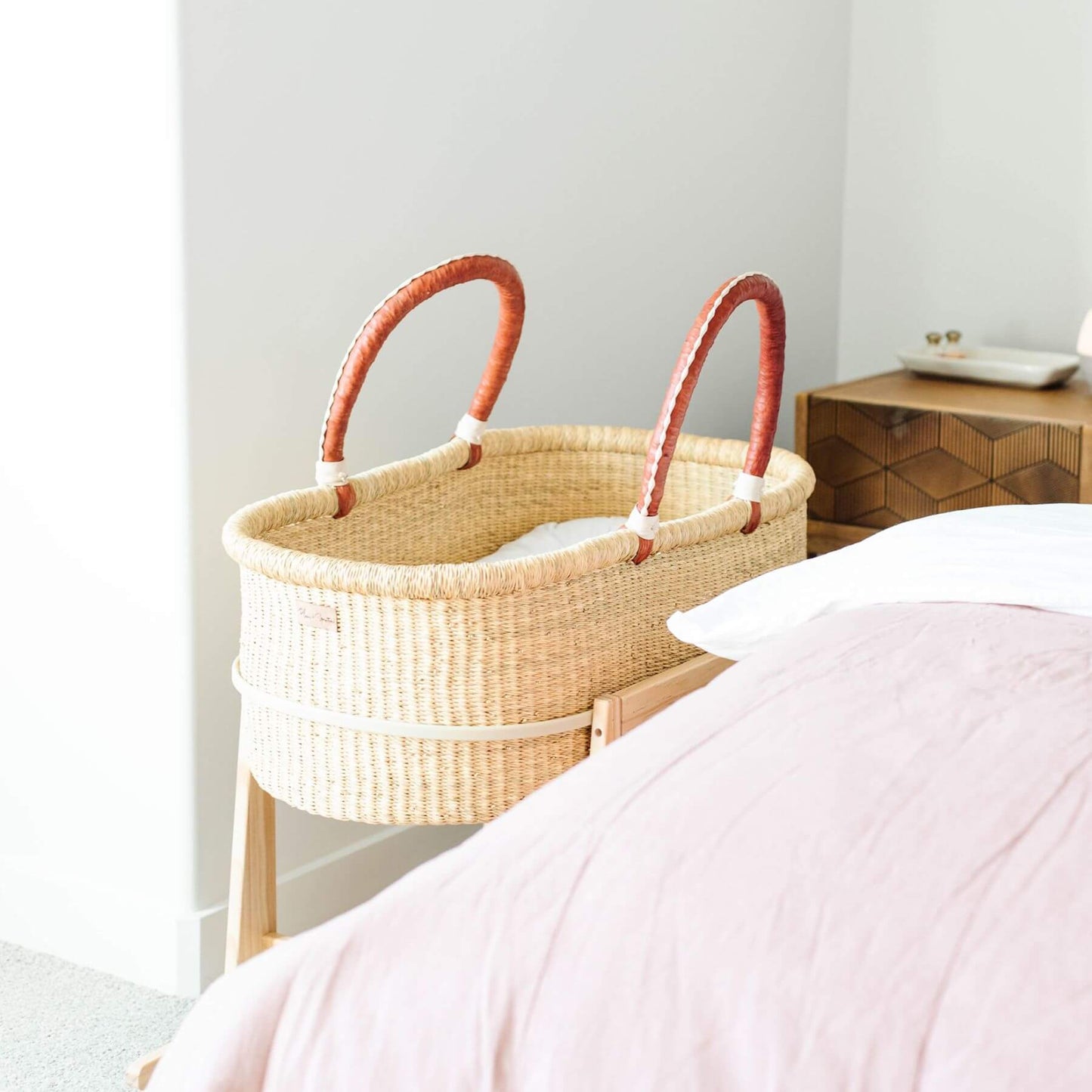 Plum+Sparrow Original Rocking Moses Bassinet Stand with brake Natural next to a bed in a bedroom setting.