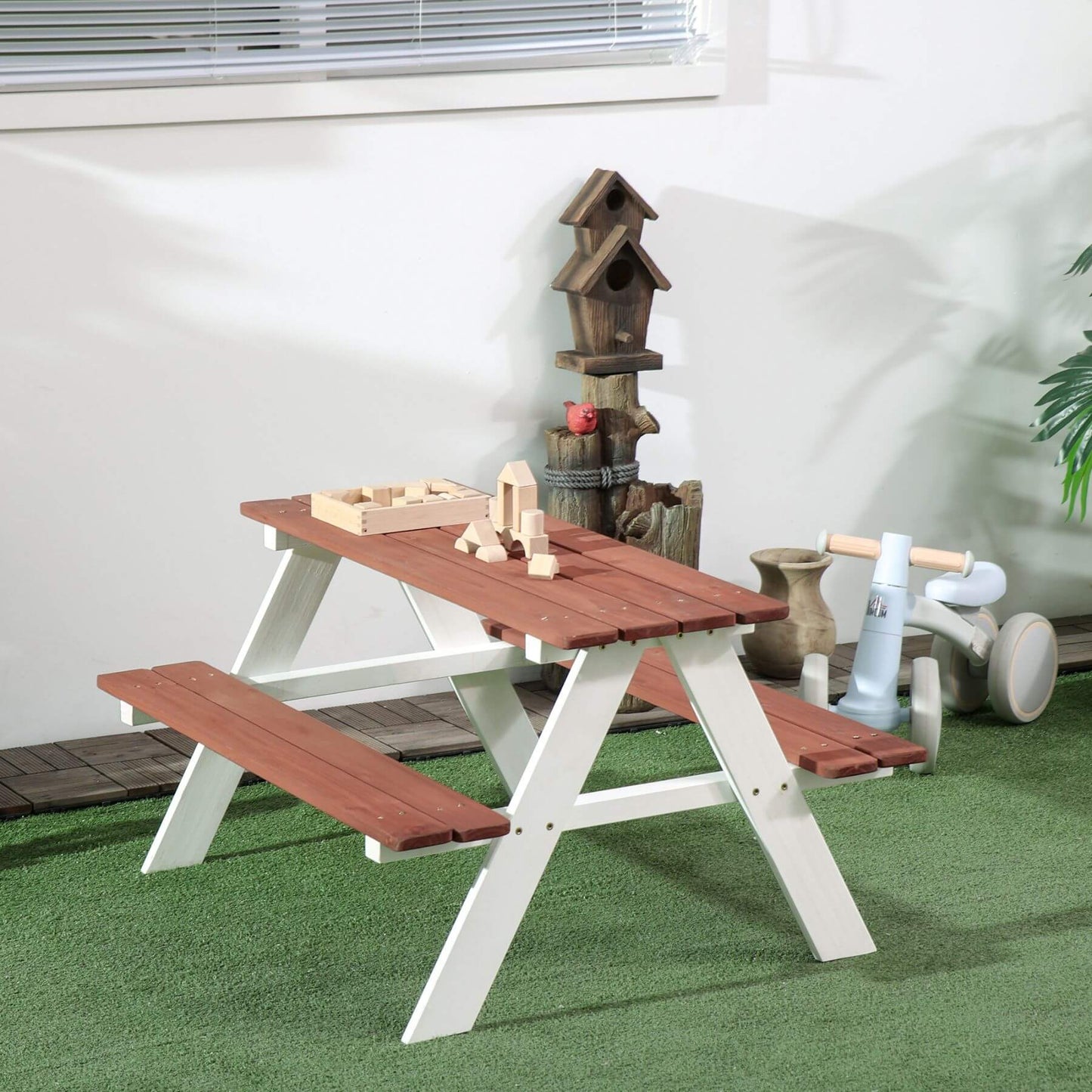 Wooden picnic table with benches on a green surface, with toys and a birdhouse in the background.