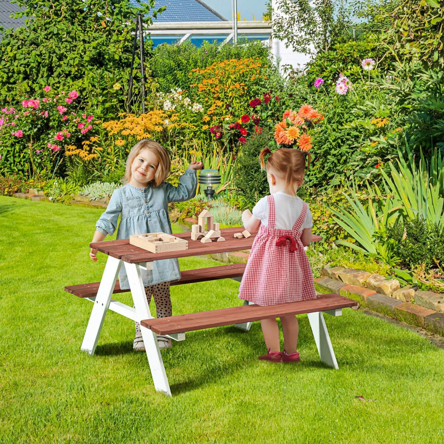 Two children playing at a wooden picnic table in a garden with flowers and greenery.