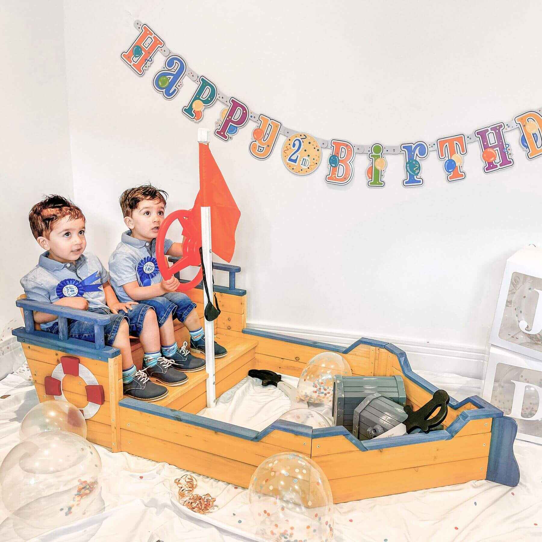 Two children sitting on a toy pirate ship with a 'Happy Birthday' banner above.