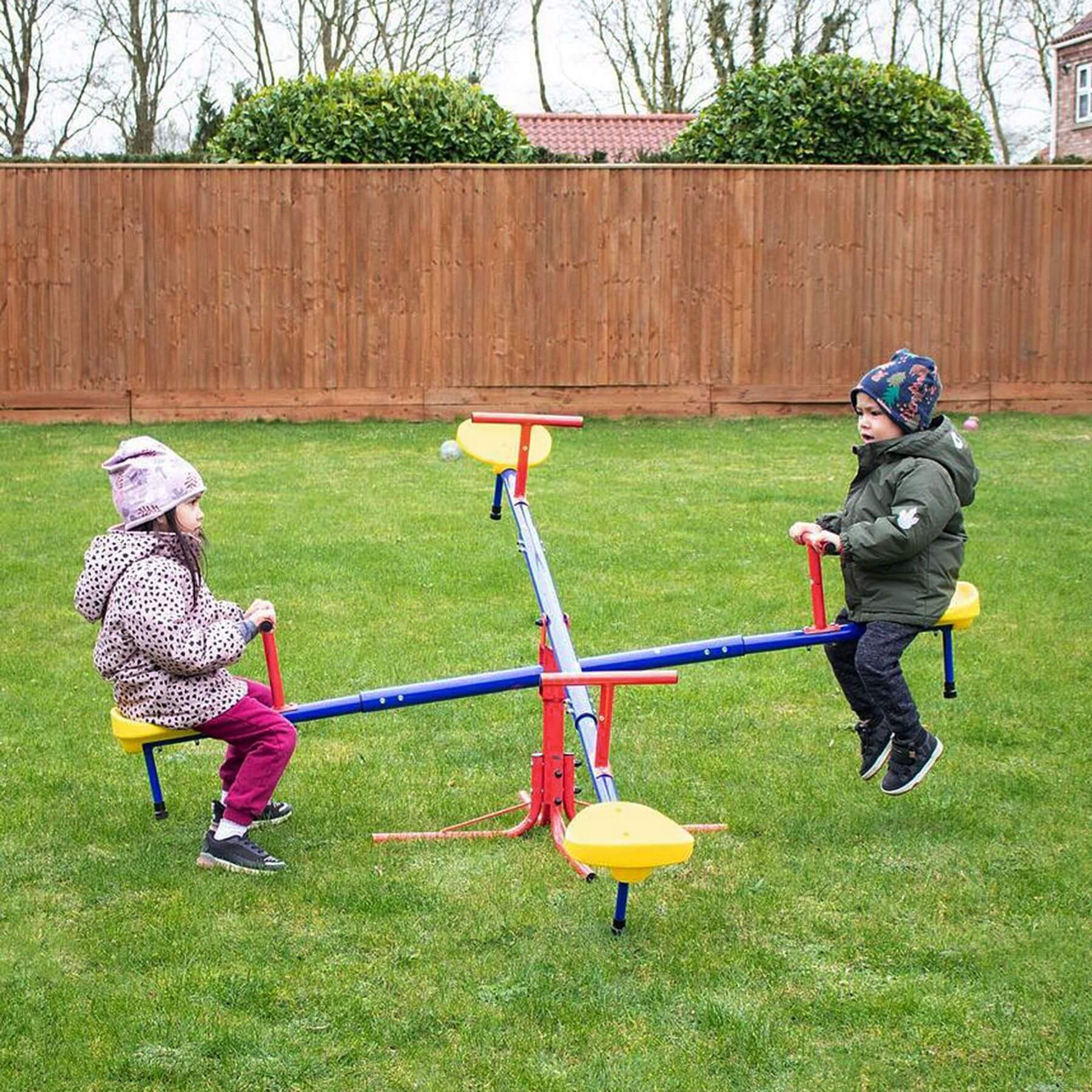 Two children playing on a seesaw in a backyard with a wooden fence and trees in the background.