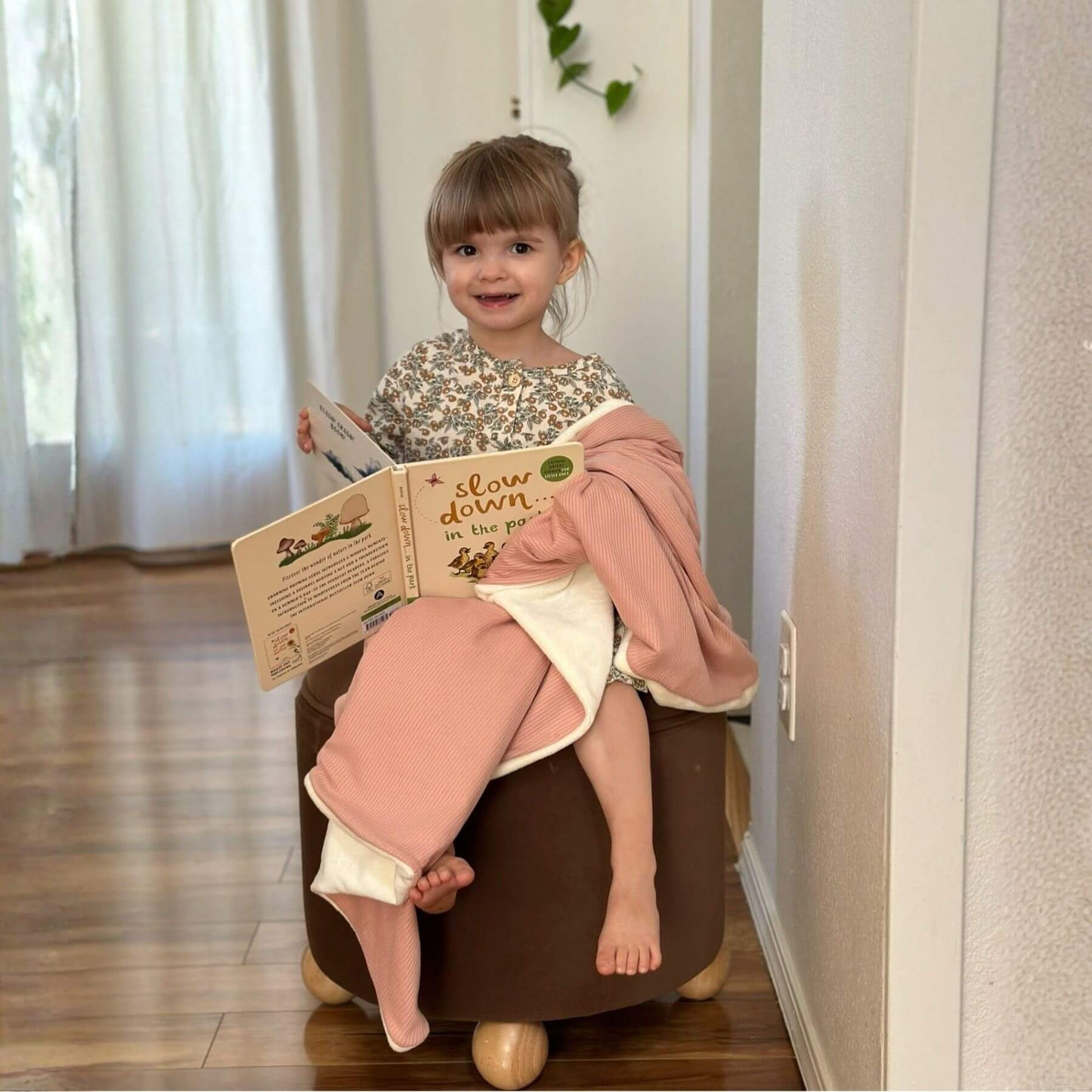 Child sitting on a brown ottoman, wrapped by Little Mae & Co Peony Ribbed Toddler Blanket, reading a book in a home setting.