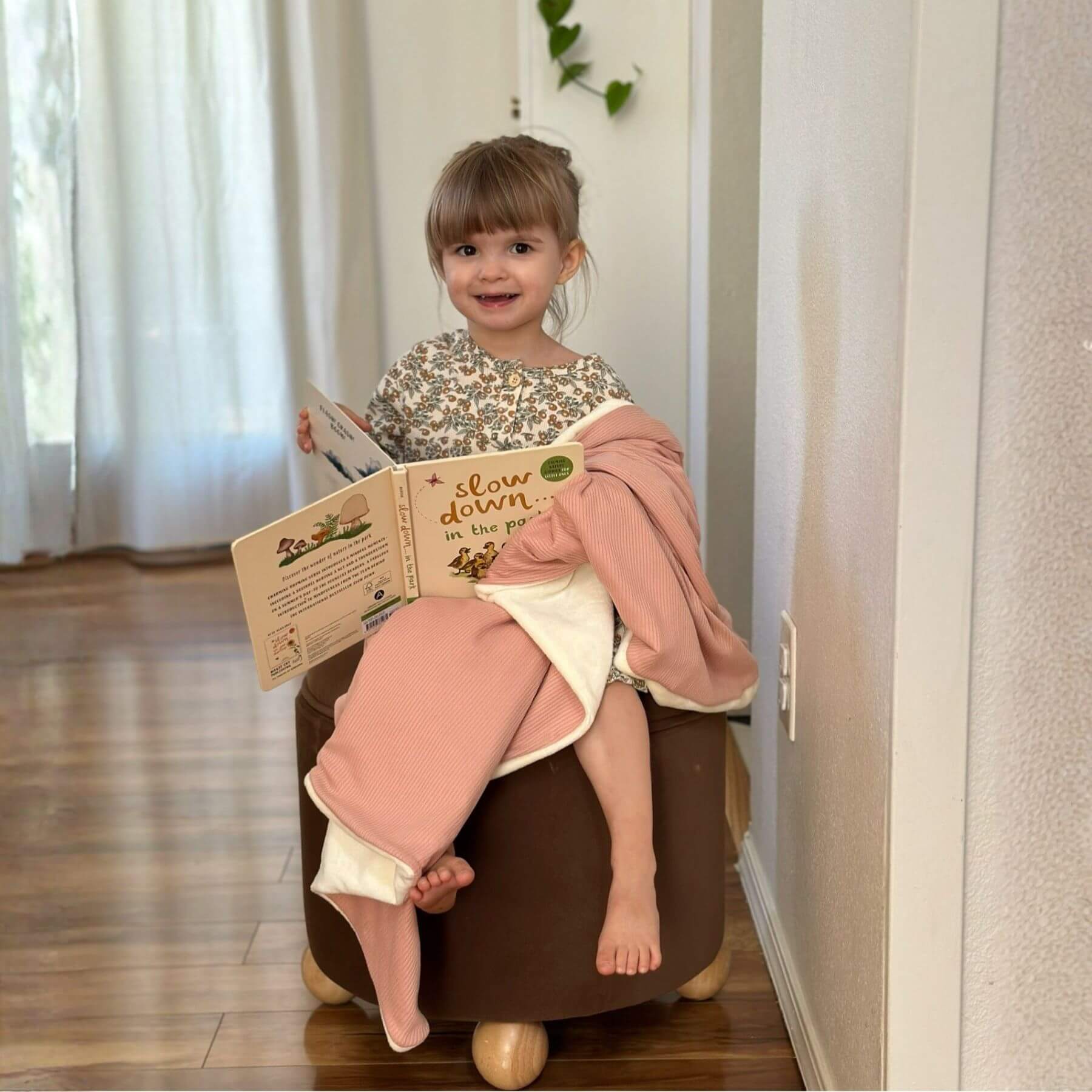 Child sitting on a brown ottoman, wrapped by Little Mae & Co Peony Ribbed Toddler Blanket, reading a book in a home setting.