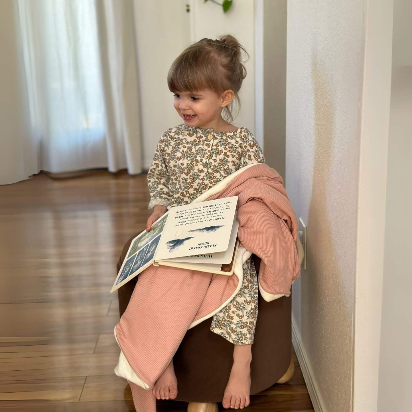Child sitting on a chair, wrapped by Little Mae & Co Peony Ribbed Toddler Blanket, holding a book in a room with white curtains.