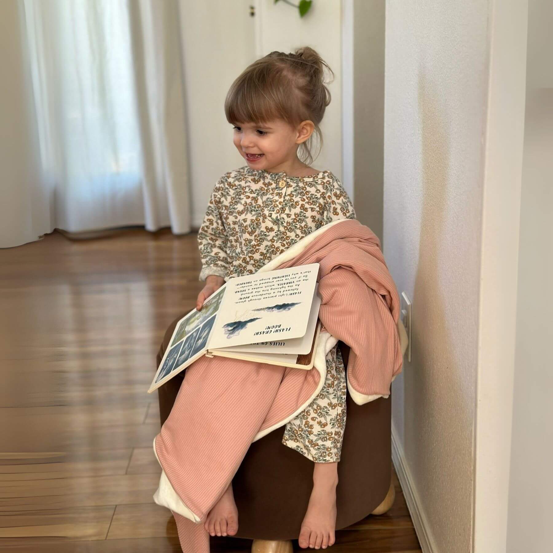 Child sitting on a chair, wrapped by Little Mae & Co Peony Ribbed Toddler Blanket, holding a book in a room with white curtains.