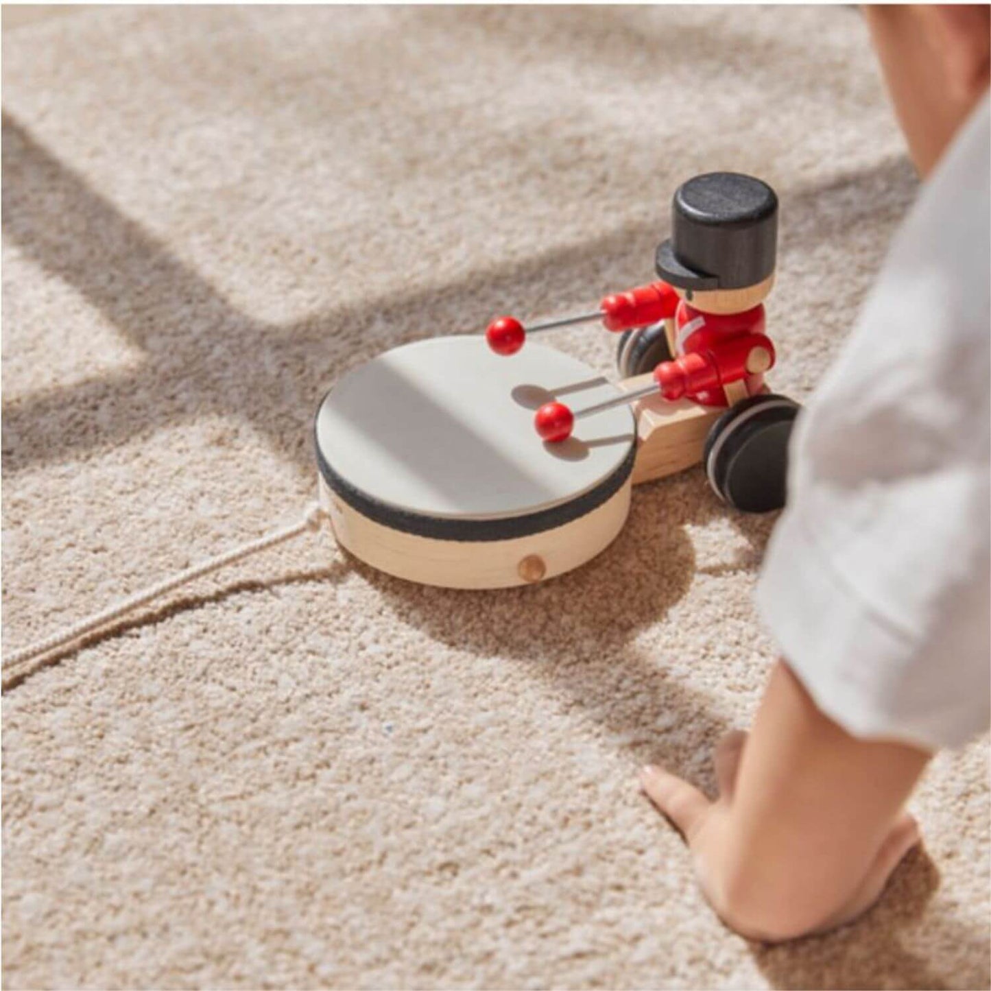 A child's hand and arm are visible as they play with PlanToys Pull Along Drummer on a beige carpet.