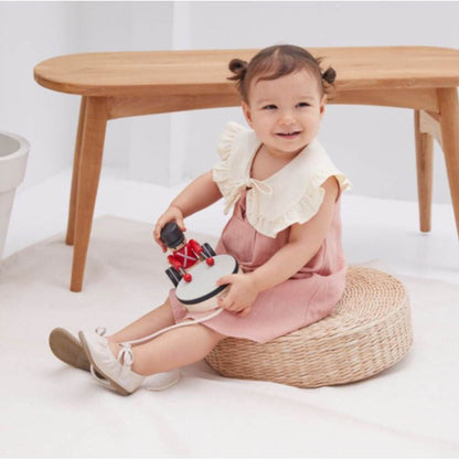 A toddler girl sits on a round woven floor cushion in front of a light wood table, holding PlanToys Pull Along Drummer