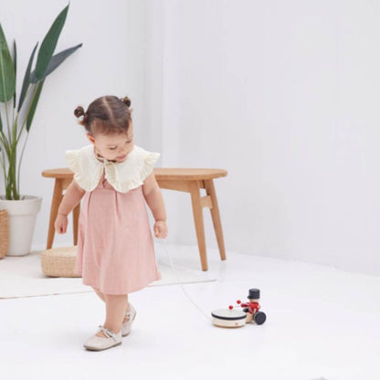 A toddler girl in a pink dress pulling PlanToys Pull Along Drummer on a white floor