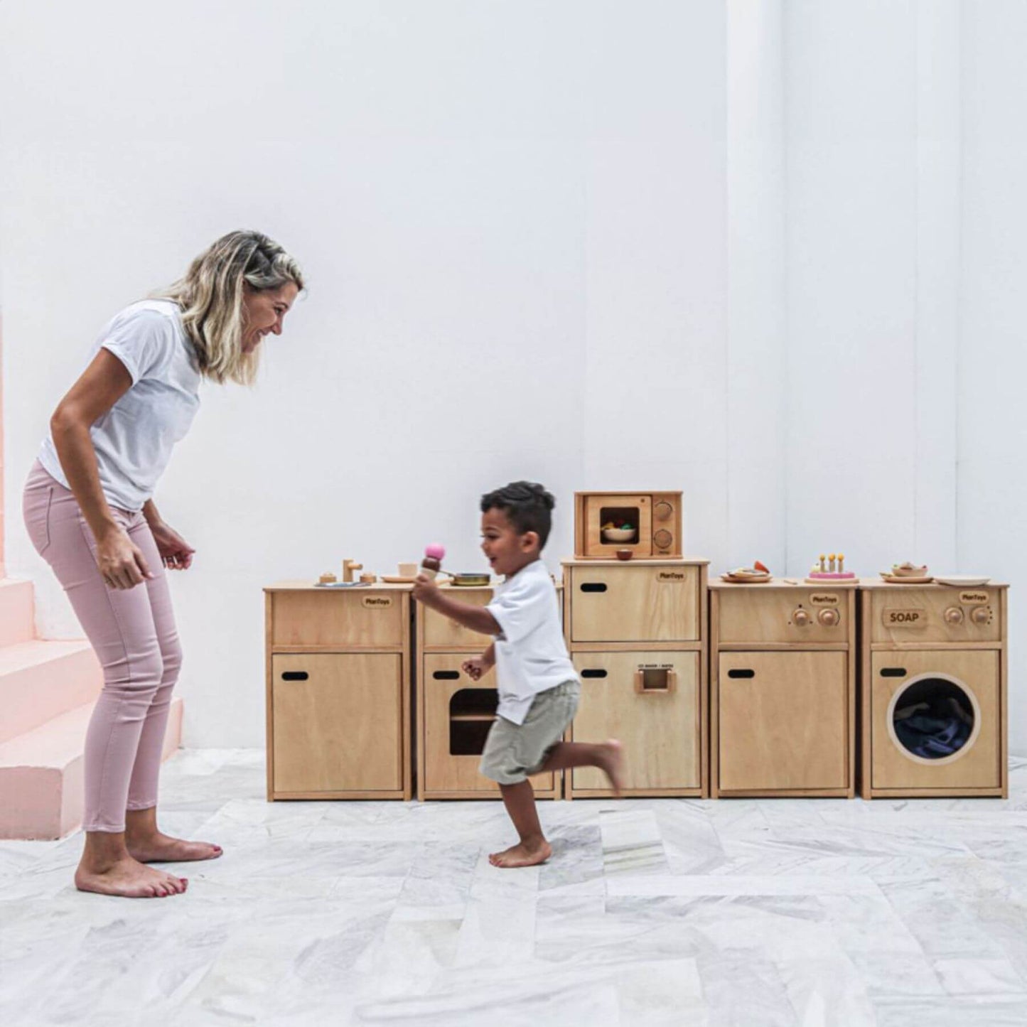 Woman and child playing with wooden toy kitchen set on a white floor.