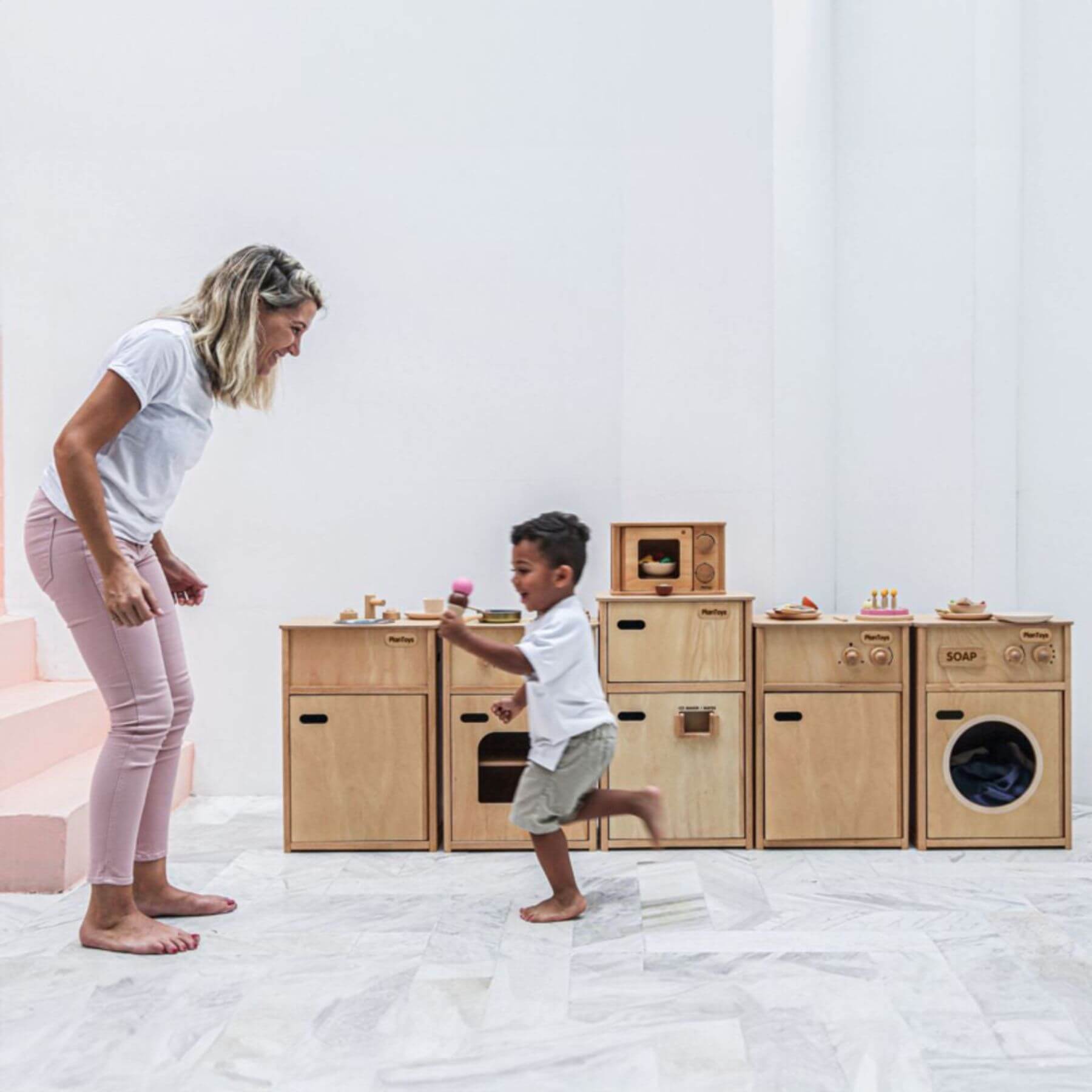 Woman and child playing with wooden toy kitchen set on a white floor.