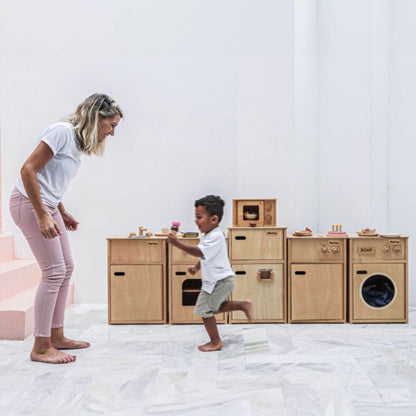 Woman and child playing with wooden toy kitchen set on a white floor.