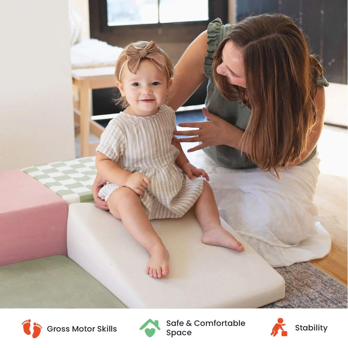A toddler sits on Tiny Land® PlayNest Climbing Blocks - Morandi while a woman kneads their legs in a bright indoor room.