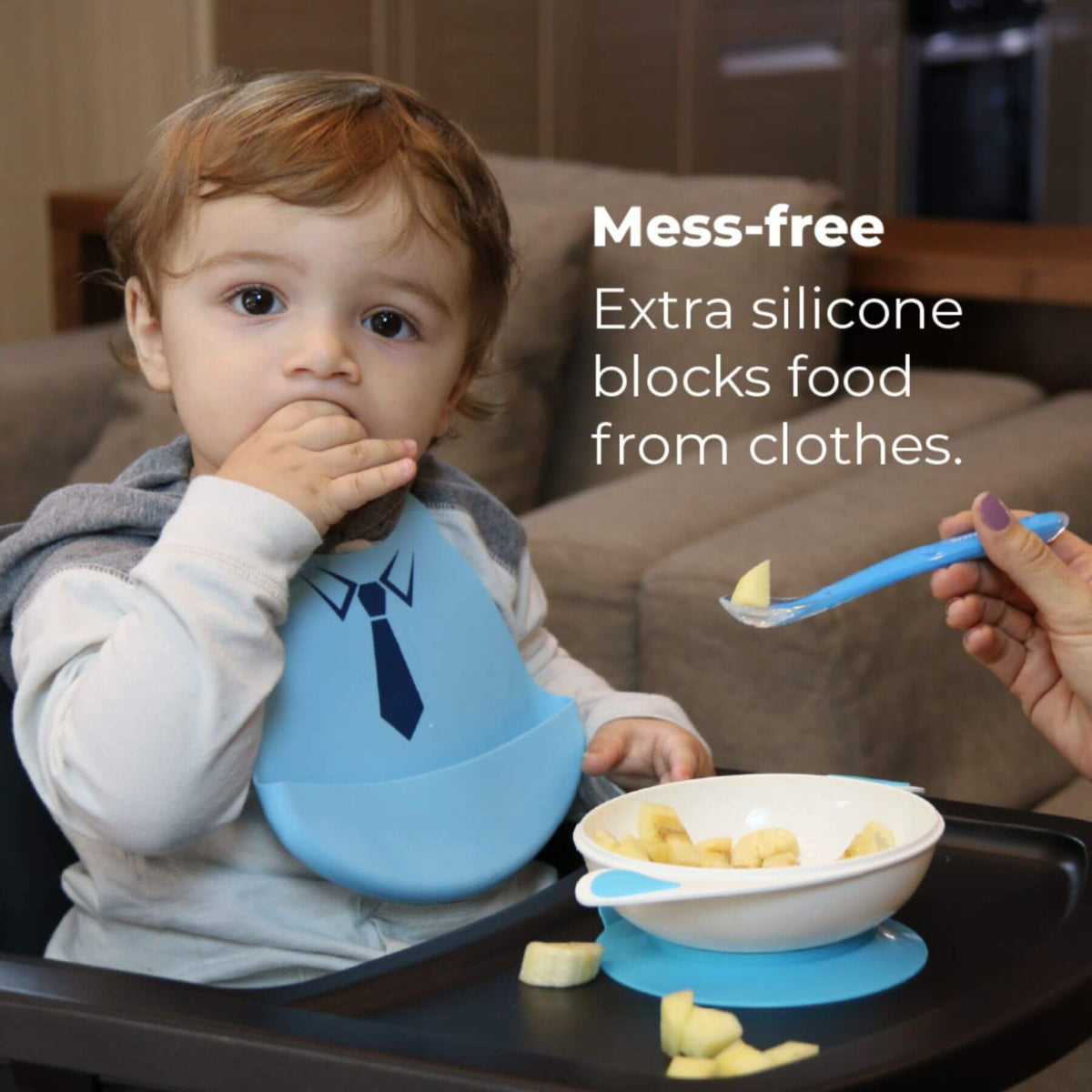 A baby seated in a high chair wearing a light blue bib with a dark blue tie graphic, eating from a white bowl with a blue silicone base on a black tray. A hand holds a blue spoon offering food.