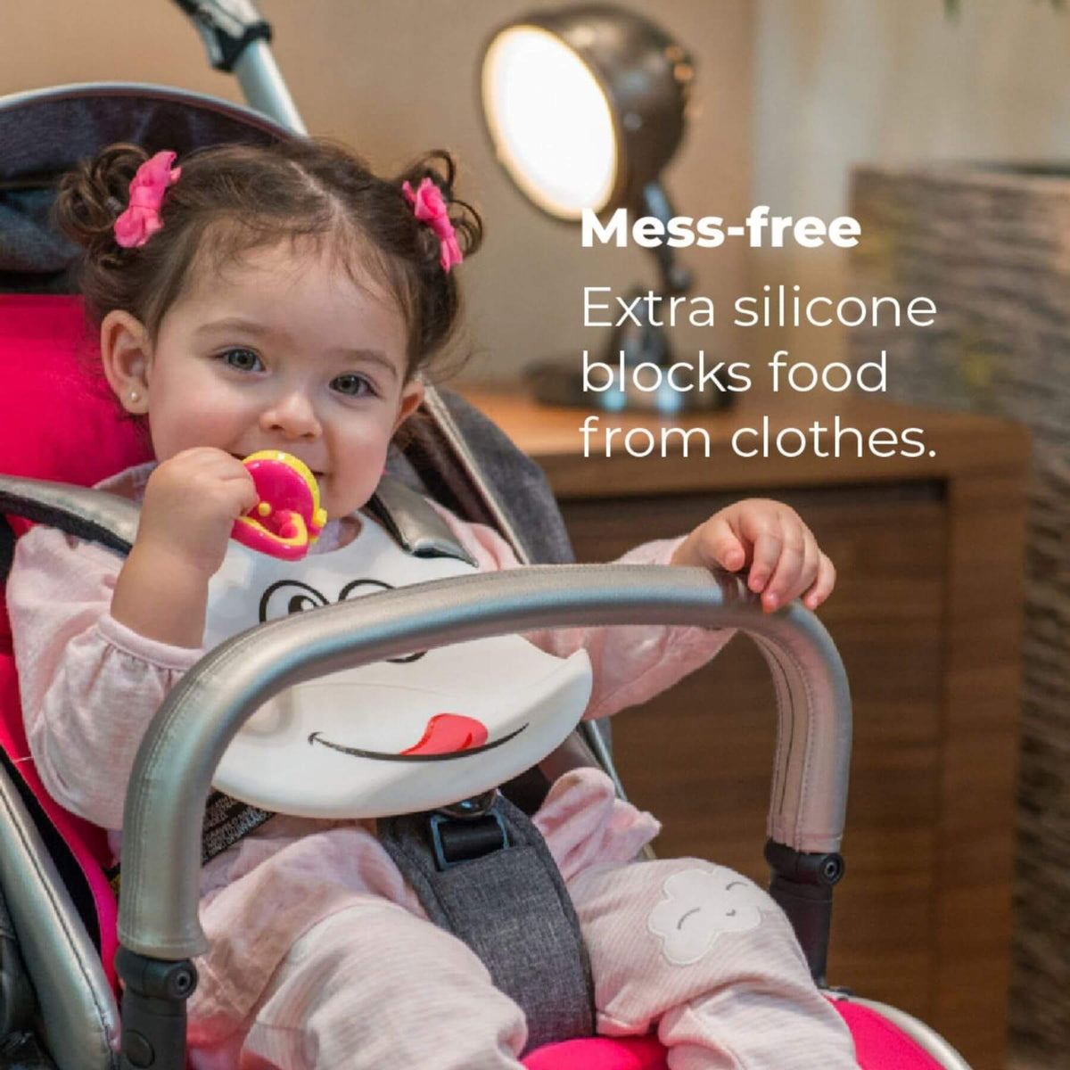 A baby in a pink stroller wearing a white bib with a cartoon face, holding a pink and yellow teething toy. White text on the right reads 'Mess-free Extra silicone blocks food from clothes.