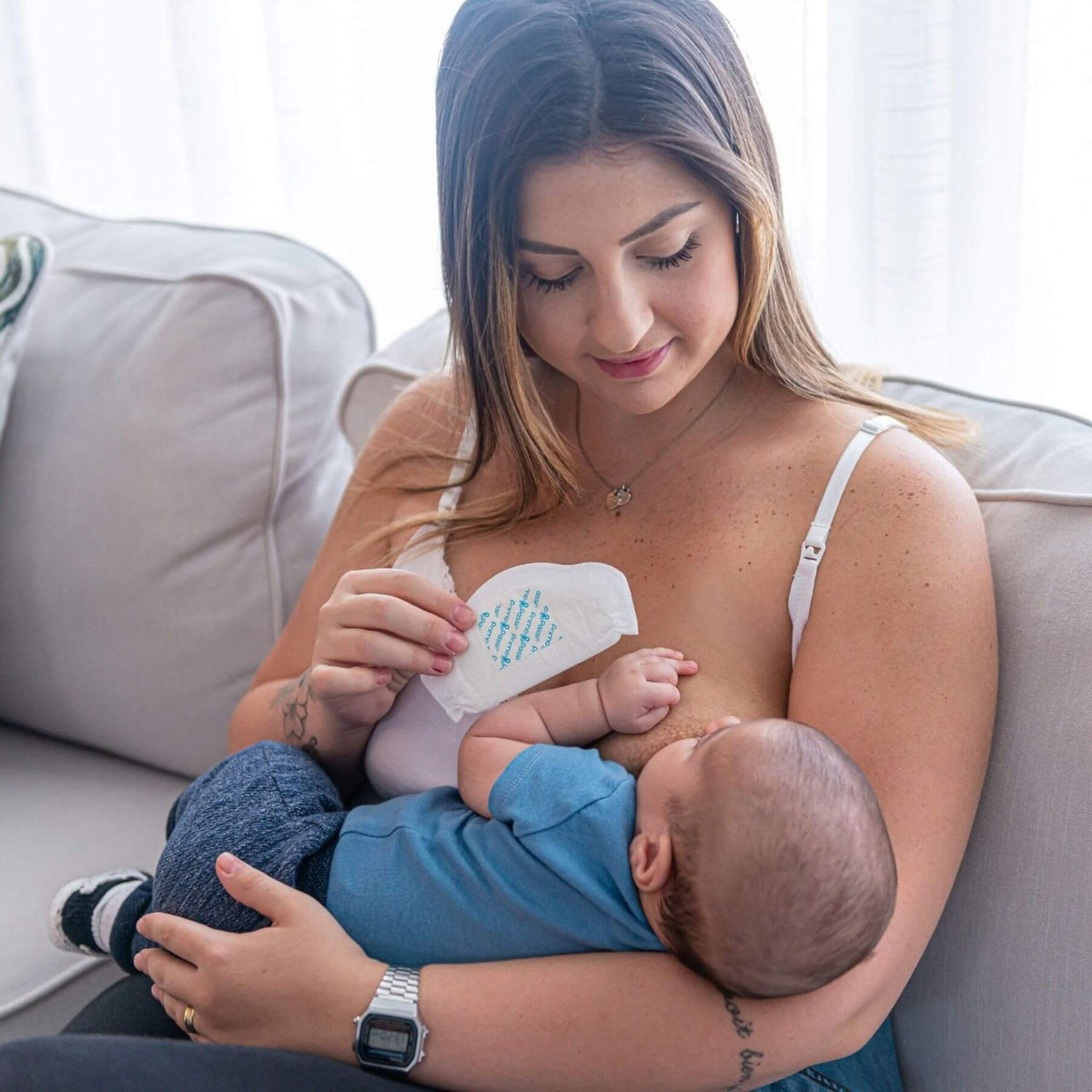 A woman breastfeeding a baby while holding a Primo Passi Disposable Nursing Pad, sitting on a light gray couch in a bright room.