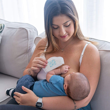 A woman breastfeeding a baby while holding a Primo Passi Disposable Nursing Pad, sitting on a light gray couch in a bright room.