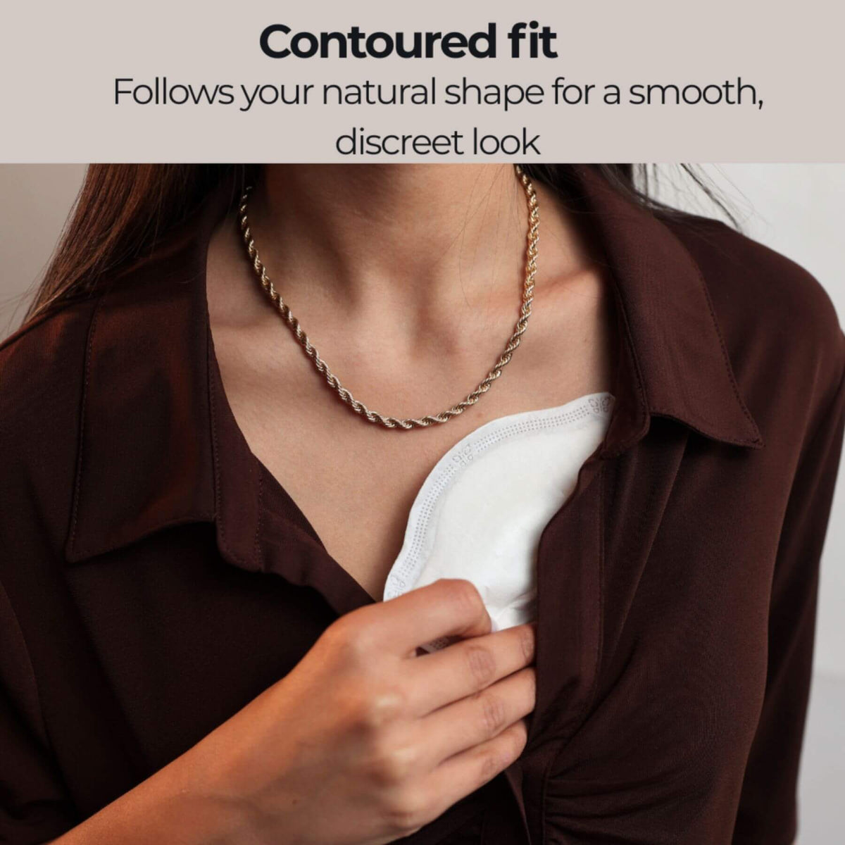 A close-up of a woman wearing a bronze rope chain necklace over a Primo Passi Disposable Nursing Pad and a dark brown collared blouse. Text at the top reads 'Contoured fit Follows your natural shape for a smooth, discreet look'.