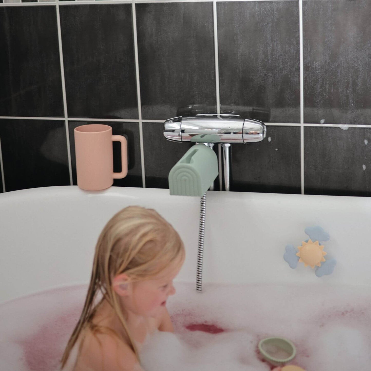 Child in a bubble bath with a pink mug and green toy, against a black tiled bathroom wall.