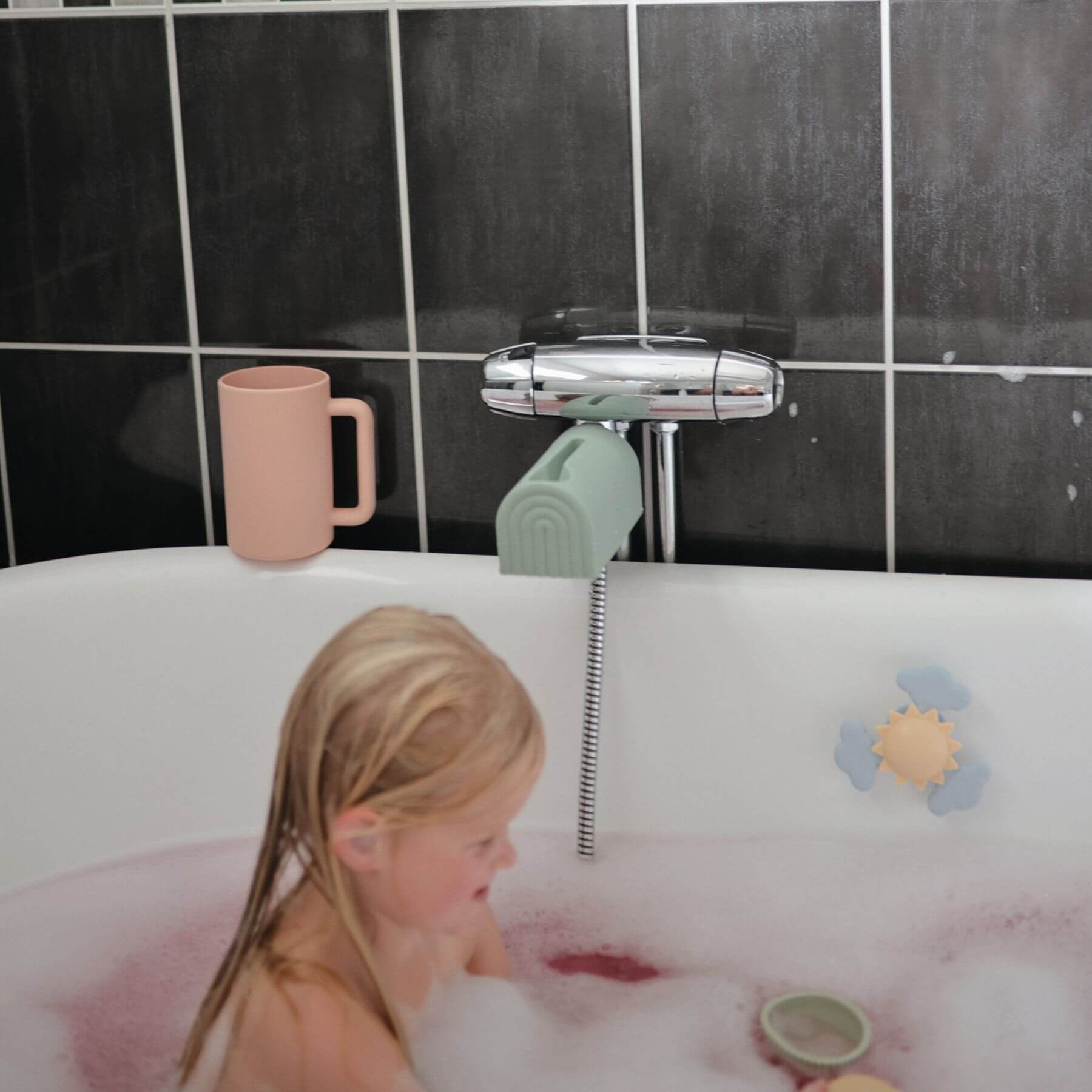 Child in a bubble bath with a pink mug and green toy, against a black tiled bathroom wall.
