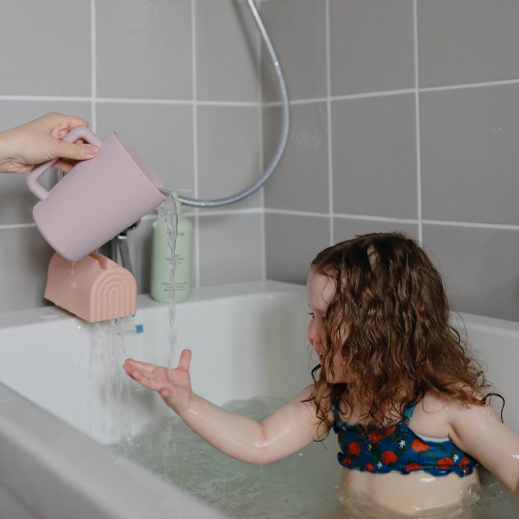 Child in a bathtub with a pink cup pouring water, surrounded by tiled walls.