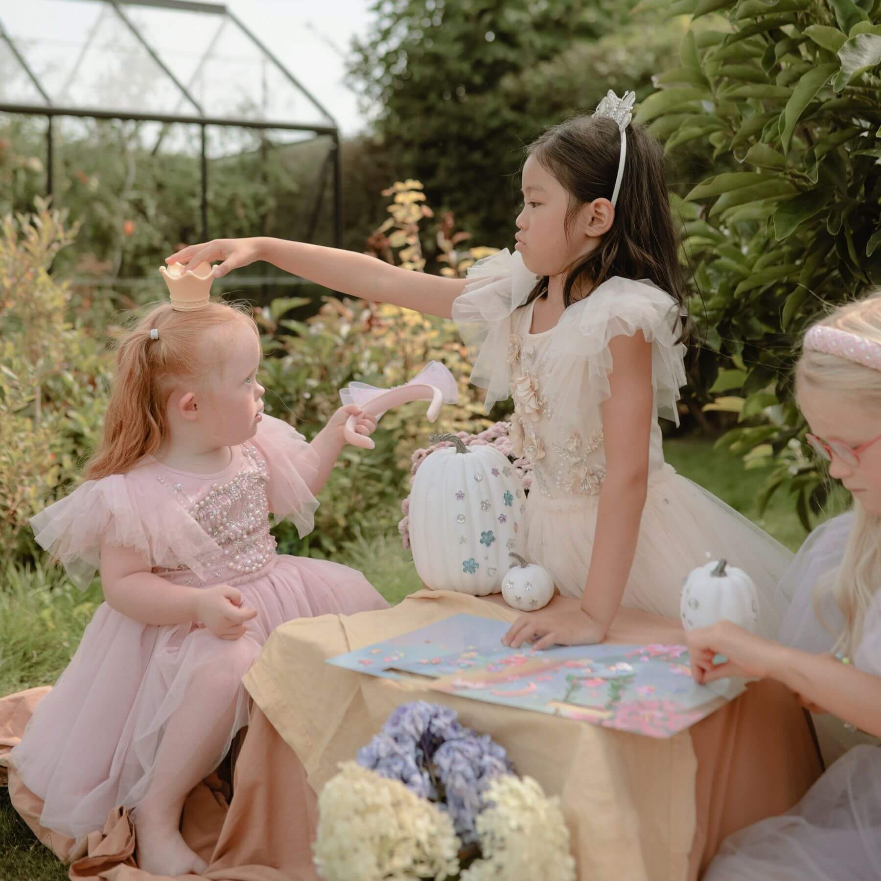 Three young girls in dresses sitting at a table outdoors with flowers and toys.