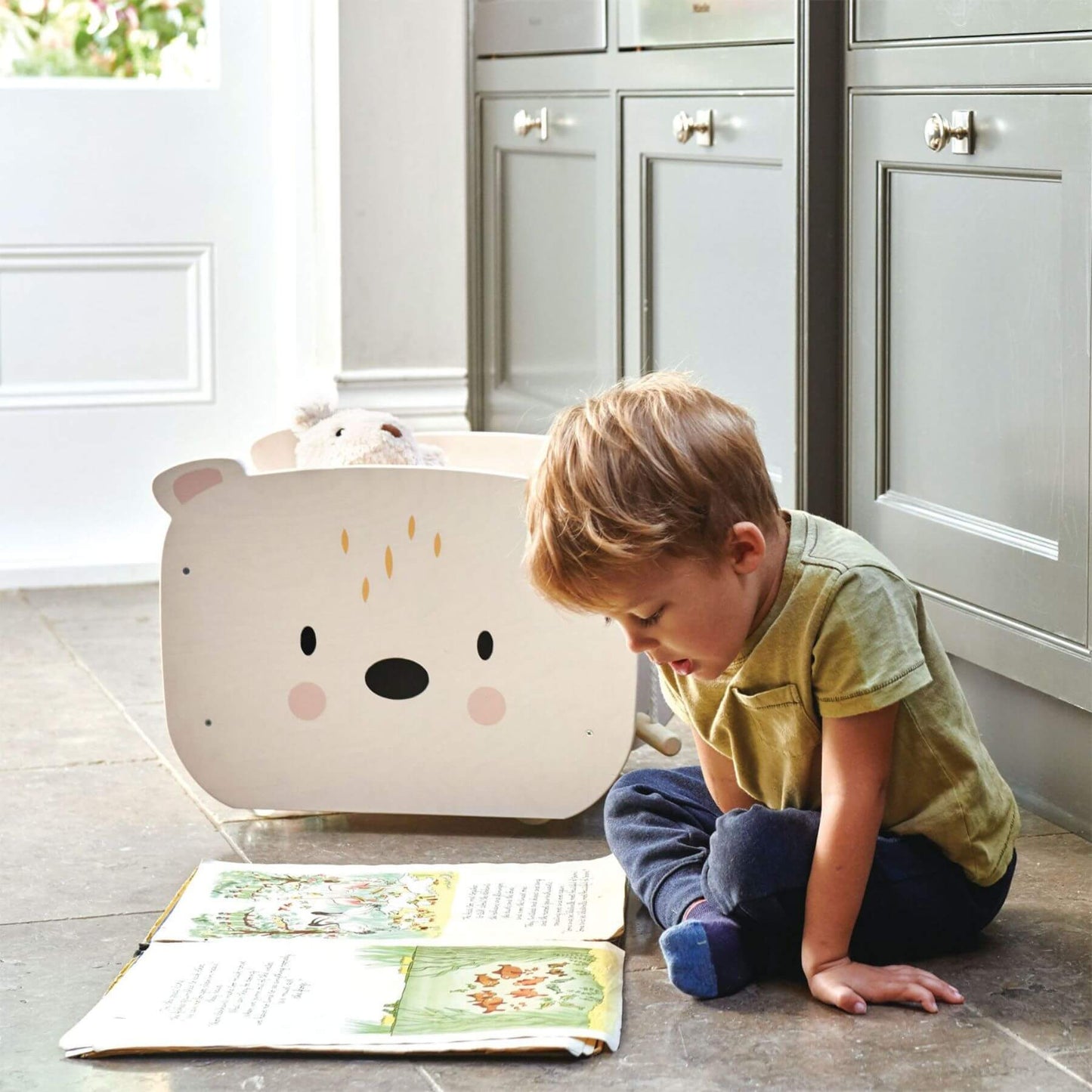 Child reading a book next to Tender Leaf Pull Along Bear Cart in a kitchen.