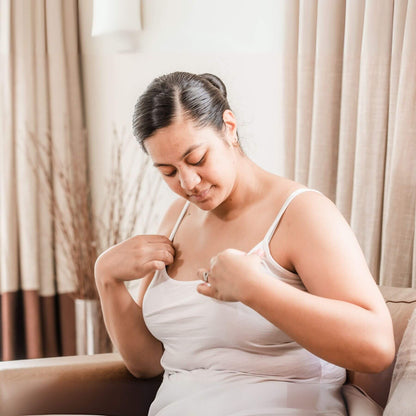 Woman adjusting a white tank top in a softly lit room with curtains.