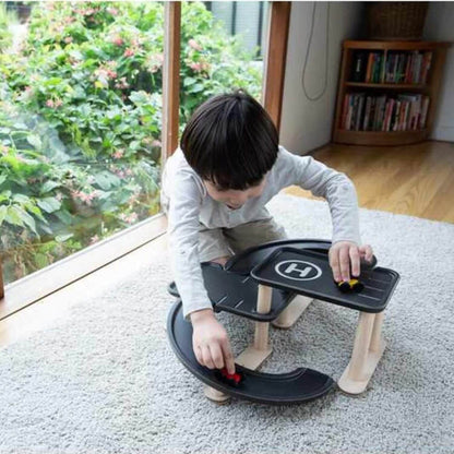 A child kneeling on a light gray carpet in a bright room, playing with PlanToys Race N Play Parking Garage
