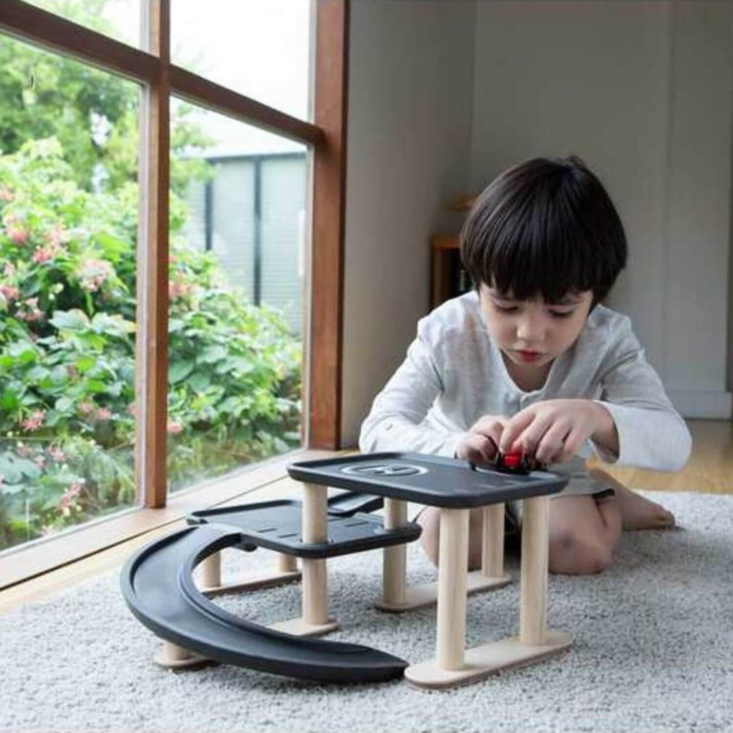A young child sits on a carpeted floor next to a large window, playing with PlanToys Race N Play Parking Garage