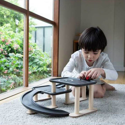 A young child sits on a carpeted floor next to a large window, playing with PlanToys Race N Play Parking Garage
