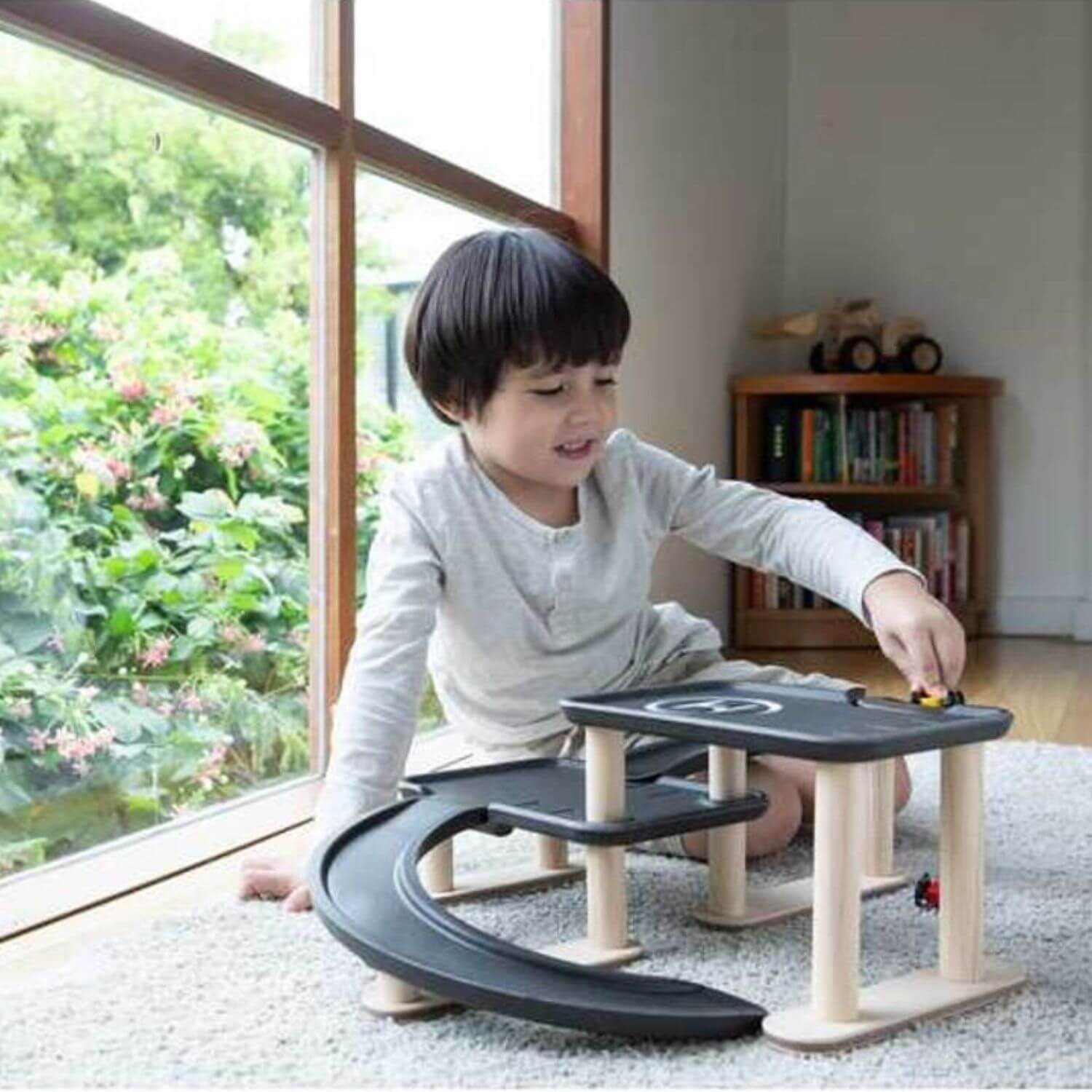 A young child sits on a carpeted floor in a bright room, playing with aPlanToys Race N Play Parking Garage
