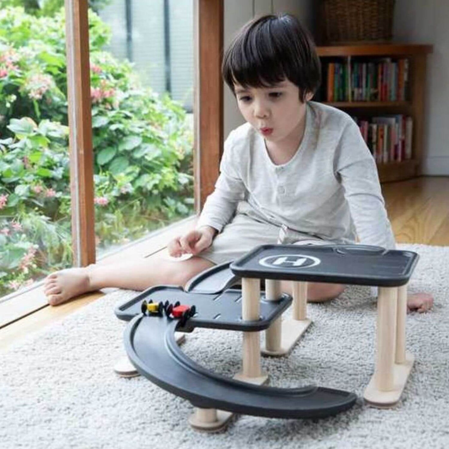 A young child sits on a light gray carpet in a bright room, playing with PlanToys Race N Play Parking Garage