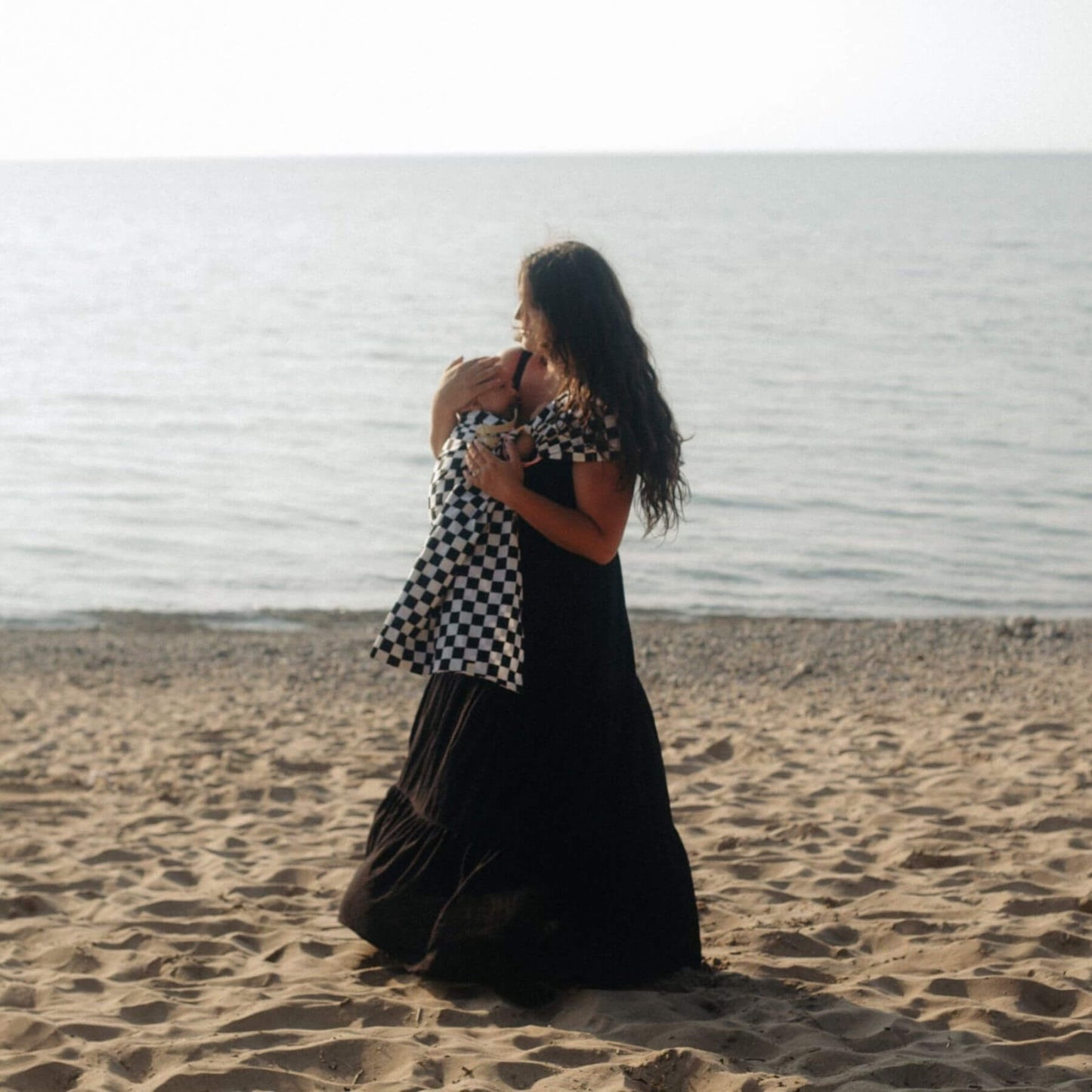 Woman in a black dress holding a baby in Little Mae & Co Race Ring Sling Carrier on a sandy beach.